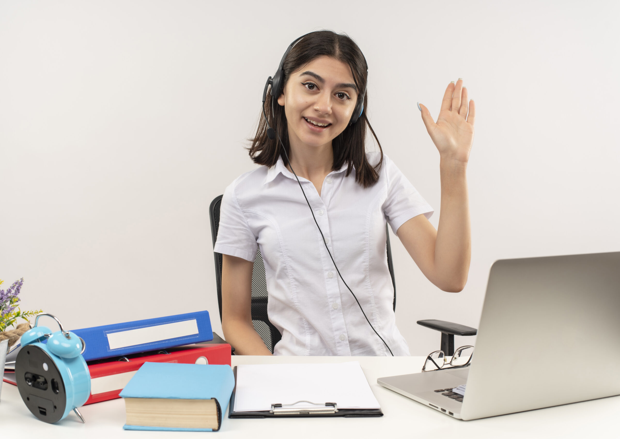 young girl in white shirt and headphones with a microphone looking at camera with smile on face waving with hand sitting at the table with folders and laptop over white background